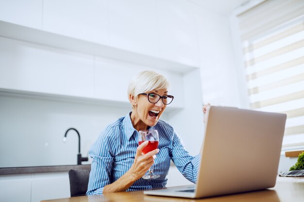 Blonde ältere Frau, die am Esstisch sitzt, Wein trinkt, Laptop betrachtet und für Leistung jubelt