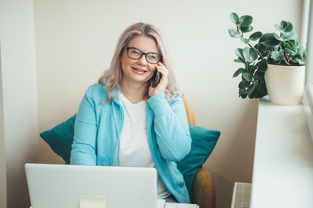 Blonde ältere Frau mit Brille, die am Telefon spricht, während sie entfernt am Computer arbeitet