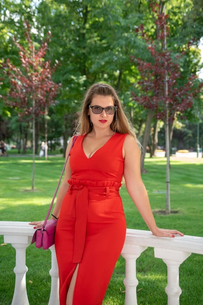 Blonde attraktive junge Dame in elegantem rotem Kleid und schwarzer Sonnenbrille auf Parkhintergrund Vertikaler Rahmen