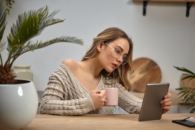 Blonde Bloggerin in beigefarbenem Strickpullover mit Brille, die rosa Tasse hält und auf das Tablet schaut In der Küche am Holztisch mit Palme im weißen Topf, Smartphone und Plastikkarte darauf sitzen Nahaufnahme