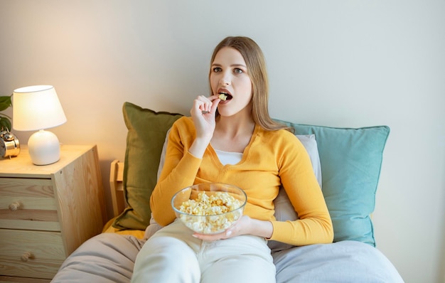 Blonde Dame schaut sich einen Film im Fernsehen an und isst Popcorn im Schlafzimmer