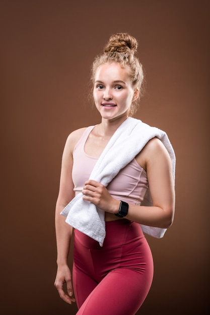 Blonde, fitte Frau in Sportkleidung, die ein weißes weiches Handtuch hält, während sie nach dem Indoor-Training auf braun isoliert eine kurze Pause macht
