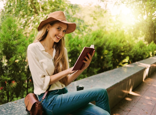 Blonde Frau des lässigen Stils im Filzhut, der ein Buch draußen in einem Stadtpark liest