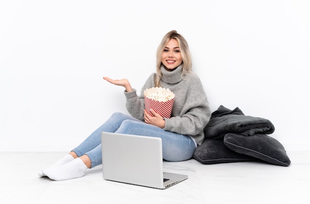 Blonde Frau des Teenagers, die Popcorn isst, während sie einen Film auf dem Laptop betrachtet, der imaginären Copyspace auf der Handfläche hält, um eine Anzeige einzufügen