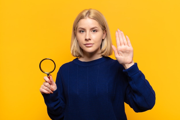 Blonde Frau, die ernst, streng, unzufrieden und wütend aussieht und offene Handfläche zeigt, die eine Stopp-Geste macht