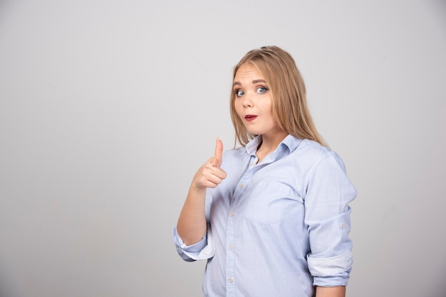 Blonde Frau, die gegen graue Wand steht und Daumen zeigt.