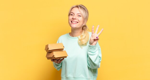 Blonde Frau, die lächelt und freundlich aussieht, die Nummer drei oder die dritte mit der Hand nach vorne zeigt und herunterzählt