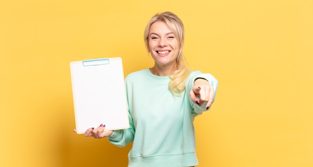 Blonde Frau, die mit einem zufriedenen, selbstbewussten, freundlichen Lächeln auf Kamera zeigt und Sie wählt