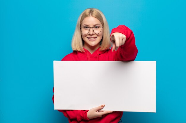 Blonde Frau, die mit einem zufriedenen, selbstbewussten, freundlichen Lächeln zeigt und Sie wählt