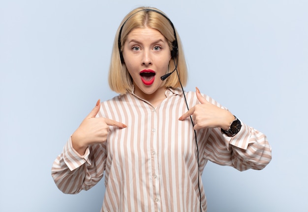 Blonde Frau, die sich glücklich, überrascht und stolz fühlt und mit einem aufgeregten, erstaunten Blick auf sich selbst zeigt