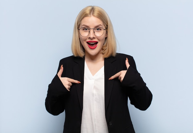 Blonde Frau, die sich glücklich, überrascht und stolz fühlt und mit einem aufgeregten, erstaunten Blick auf sich selbst zeigt