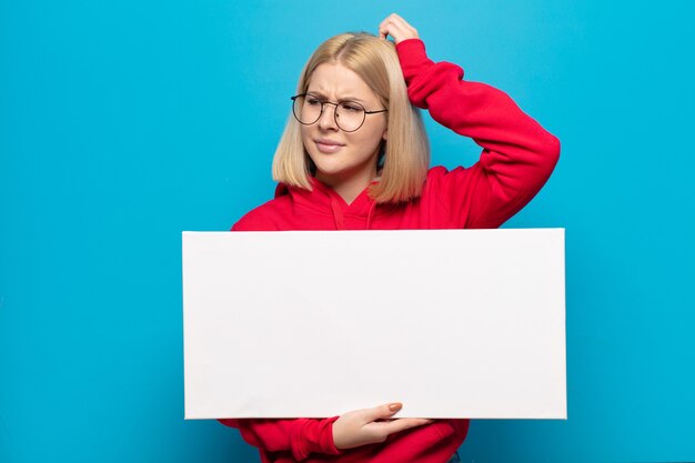 Blonde Frau, die sich verwirrt und verwirrt fühlt, sich am Kopf kratzt und zur Seite schaut