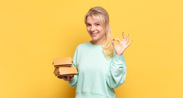 Blonde Frau fühlt sich glücklich, entspannt und zufrieden, zeigt Zustimmung mit okay Geste, lächelnd