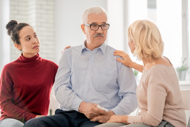 Blonde Frau im Alter und Mädchen gemischter Abstammung beruhigen älteren Gruppenmitglied oder Patienten während der Psychotherapie-Sitzung