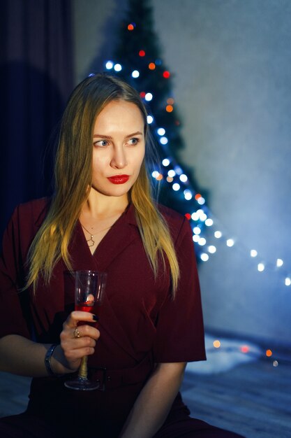 Blonde Frau im roten Kleid mit Glas Weißwein oder Champagner im Haus. Weihnachtsbaum. Neujahrsdekorationen