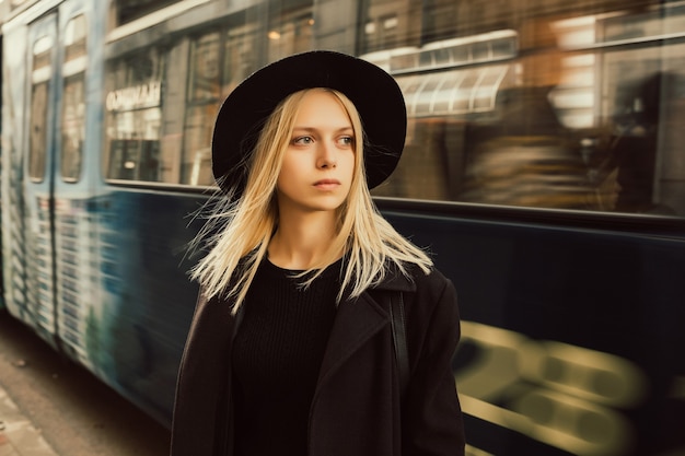 Blonde Frau im stilvollen schwarzen Mantel und im Hut, die zur Arbeit gehen