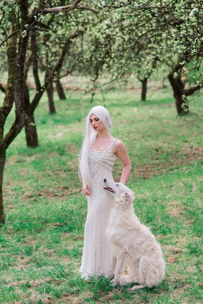 Blonde Frau im weißen Kleid, das mit ihrem russischen Wolfshundhund im Garten spielt.