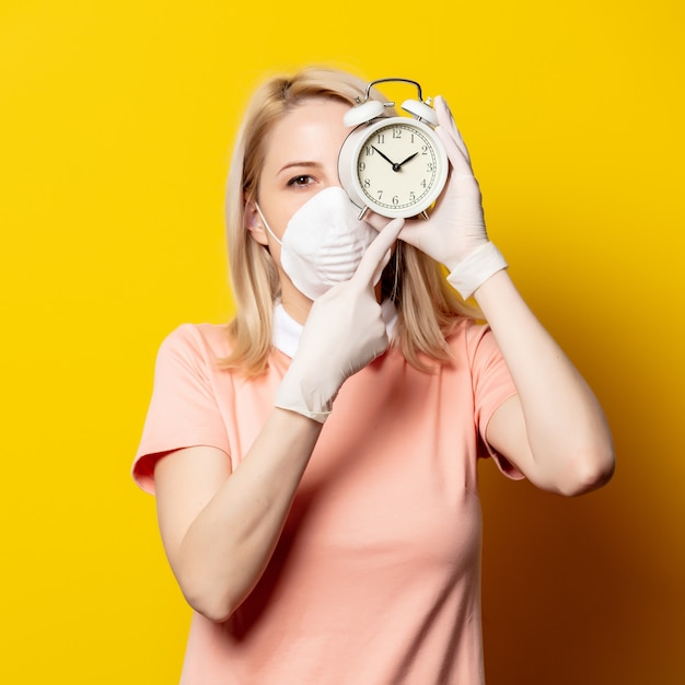 Blonde Frau in Gesichtsmaske mit Wecker auf gelber Wand