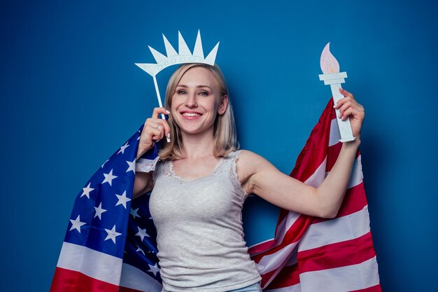 Blonde Frau in Jeans mit amerikanischer Flagge mit Papierkrone und Fackel Freiheitsstatue auf blauem Hintergrund im Studio .4. Juli Unabhängigkeitstag Feierkonzept