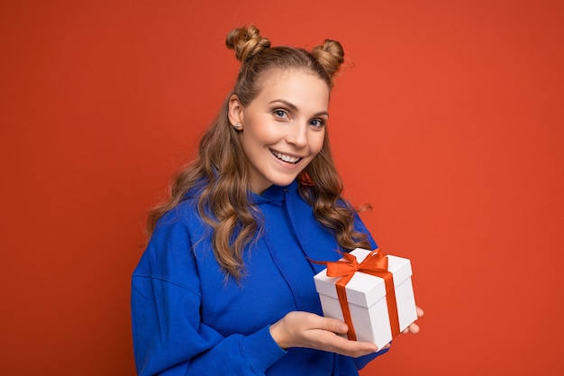 blonde Frau lokalisiert über rote Hintergrundwand, die blauen trendigen Kapuzenpulli hält, der Geschenkbox hält und Kamera betrachtet.