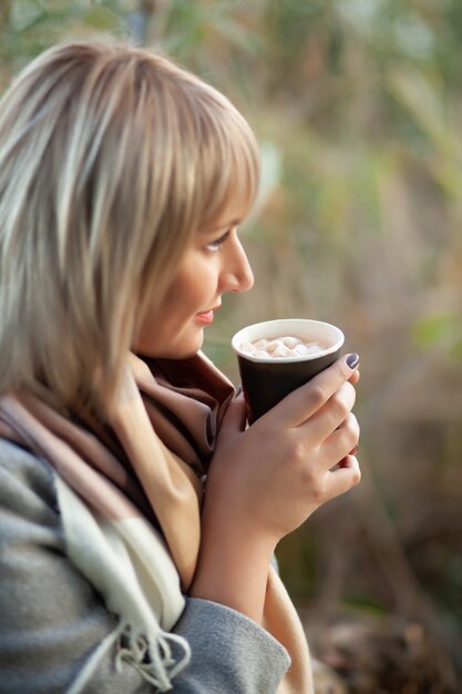 Blonde Frau mit kurzem Haarschnitt in grauem Wollmantel und kariertem Schal trinkt Kaffee-Cappuccino mit Marshmallows.