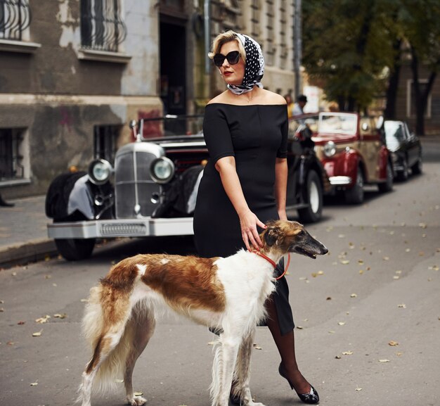 Blonde Frau mit Sonnenbrille und schwarzem Kleid in der Nähe eines alten Oldtimers mit ihrem Hund.