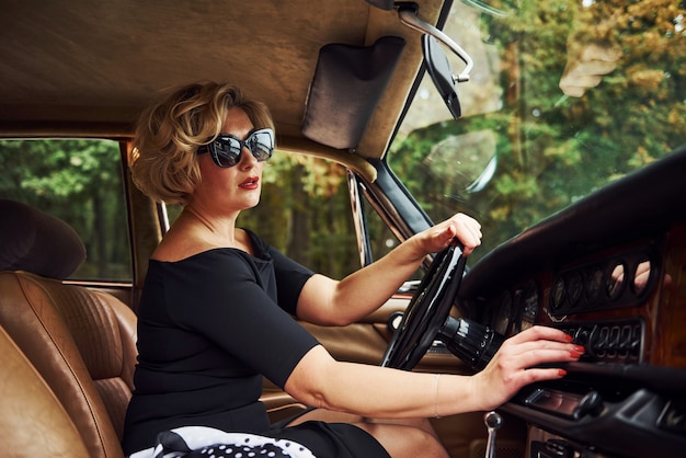 Blonde Frau mit Sonnenbrille und schwarzem Kleid sitzt in einem alten Oldtimer.