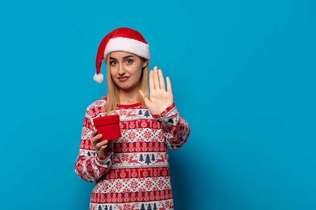 Blonde Frau mit Weihnachtsmütze, die ernst, streng, unzufrieden und wütend aussieht und offene Handfläche zeigt, die Stoppgeste macht