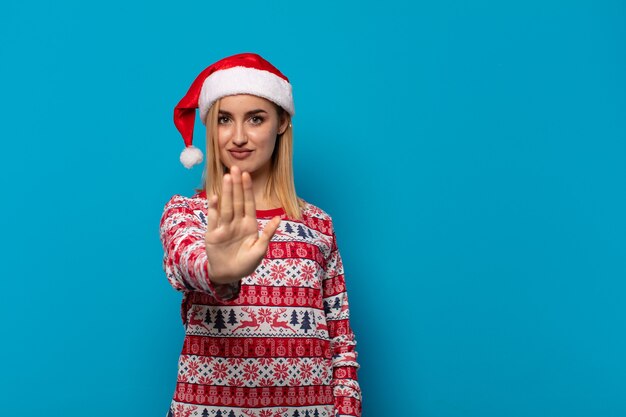 Blonde Frau mit Weihnachtsmütze, die ernst, streng, unzufrieden und wütend aussieht und offene Handfläche zeigt, die Stoppgeste macht