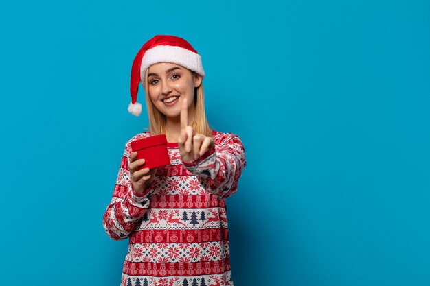 Blonde Frau mit Weihnachtsmütze, die stolz und zuversichtlich lächelt und Nummer eins triumphierend posiert und sich wie ein Anführer fühlt