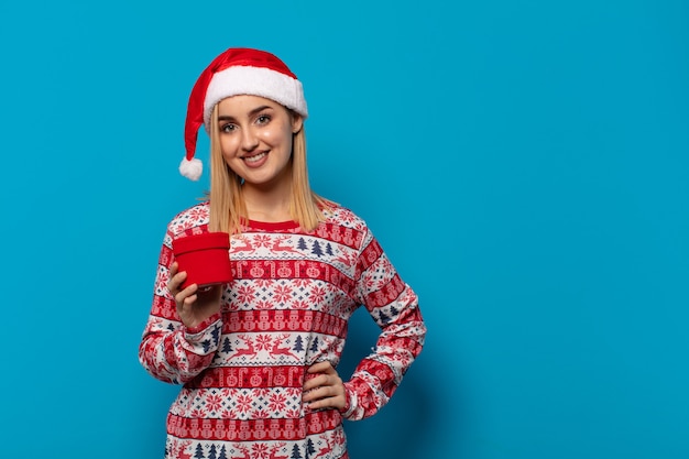 blonde Frau mit Weihnachtsmütze glücklich lächelnd mit einer Hand auf der Hüfte und selbstbewusst, positiv, stolz und freundlich