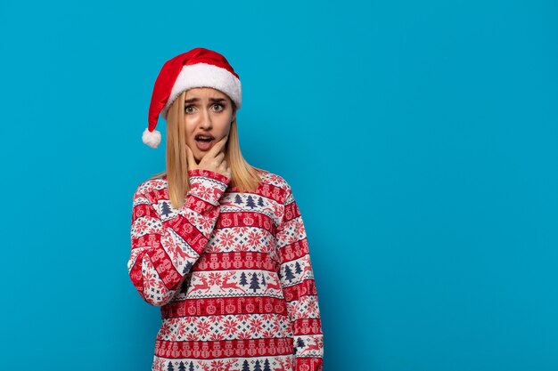 Blonde Frau mit Weihnachtsmütze mit offenem Mund und weit geöffneten Augen und Hand am Kinn, die sich unangenehm geschockt fühlt