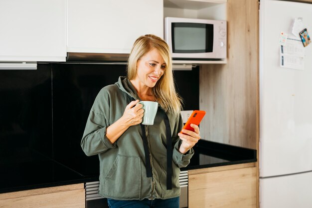 Blonde Frau mittleren Alters, die Kaffee trinkt und auf einem roten Smartphone in einer Küche SMS schreibt