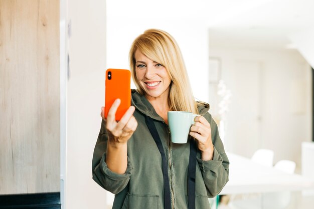 Blonde Frau mittleren Alters, die Kaffee trinkt und einen Videoanruf in einer Küche hat