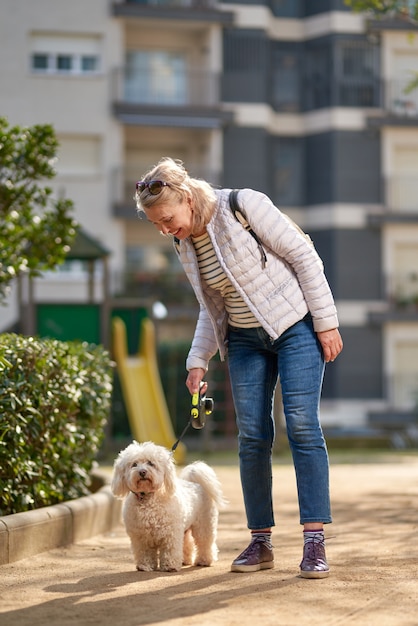 Blonde Frau mittleren Alters, die mit flauschigem weißem Hund in der Sommerstadt geht.