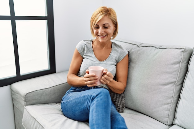 Blonde Frau mittleren Alters lächelt selbstbewusst und trinkt zu Hause Kaffee