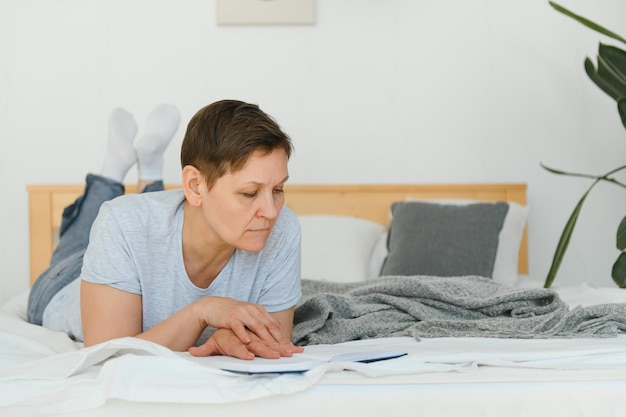 Blonde Frau mittleren Alters liest ein Buch, das zu Hause auf dem Bett liegt
