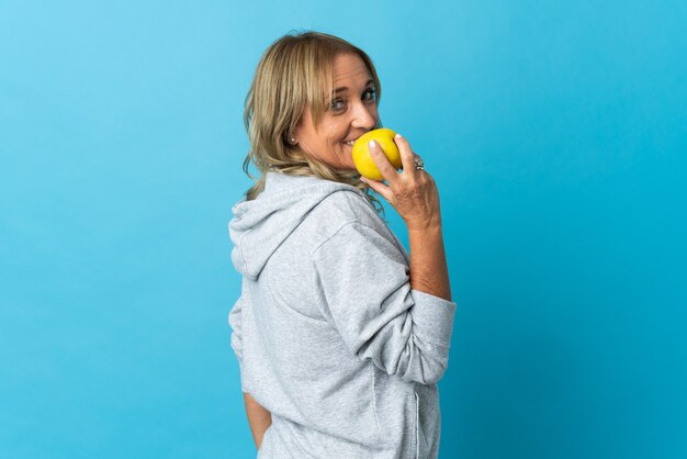 Blonde Frau mittleren Alters über isolierter Wand, die einen Apfel isst