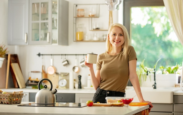 Blonde Frau steht in der Küche mit einer Tasse Kaffee