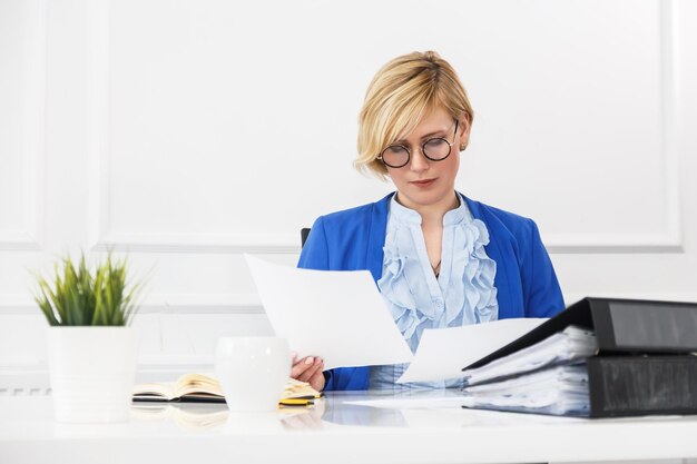 Blonde Frau trägt eine blaue Jacke, die mit Dokumenten auf dem Tisch mit einem beschäftigten Arbeitskonzept des weißen Büroraums im Blumentopf verwaltet wird