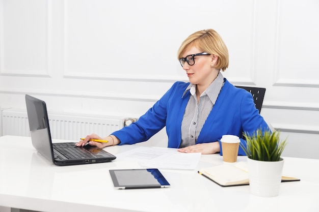 Blonde Frau trägt eine blaue Jacke, die mit Dokumenten auf dem Tisch mit einem beschäftigten Arbeitskonzept des weißen Büroraums im Blumentopf verwaltet wird