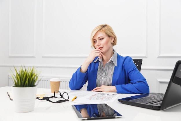 Blonde Frau trägt eine blaue Jacke, die mit Dokumenten auf dem Tisch mit einem beschäftigten Arbeitskonzept des weißen Büroraums im Blumentopf verwaltet wird