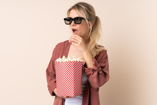 Blonde Frau über isolierter Wand mit 3D-Brille und hält einen großen Eimer Popcorn, während sie Seite schaut