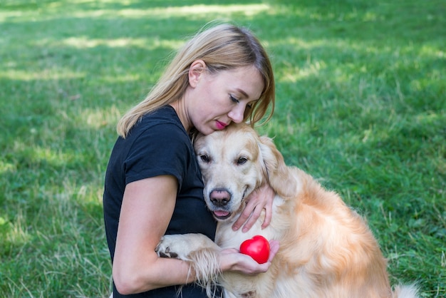 Blonde Frau umarmt Retrieverhund, der in der Hand rotes Herz hält. Das Konzept der Tierliebe.