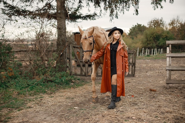Blonde Frau und braunes Pferd stehen auf einem Bauernhof. Frau mit schwarzem Kleid, rotem Ledermantel und Hut. Frau, die das Pferd berührt.