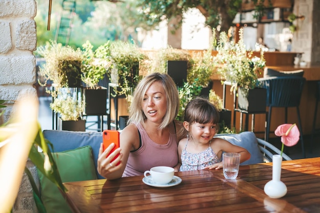 Blonde Frau und ihre kleine Tochter nutzen Smartphone für Video-Chat im Café Lifestyle-Technologie und Kommunikationskonzept