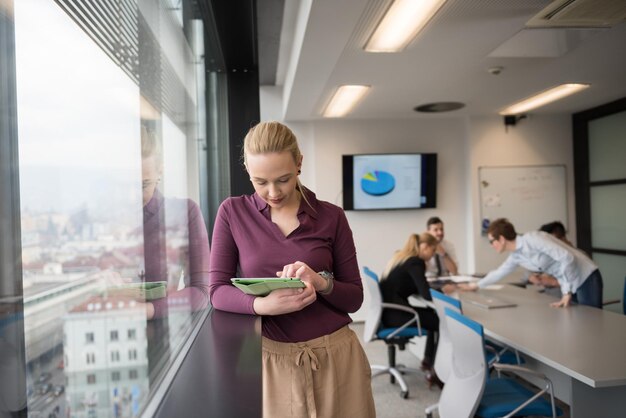 Blonde Geschäftsfrau in Freizeitkleidung, die an einem Tablet-Computer im modernen Start-Business-Büro-Interieur arbeitet. Gruppe junger Leute bei Teamsitzungen, die im Hintergrund verschwommen sind