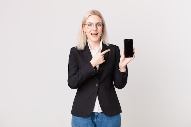 Blonde hübsche Frau, die aufgeregt und überrascht aussieht, die auf die Seite zeigt und ein Mobiltelefon hält