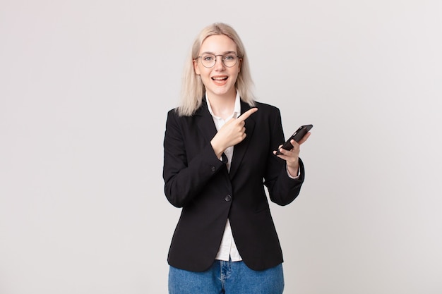 Blonde hübsche Frau, die aufgeregt und überrascht aussieht, die auf die Seite zeigt und ein Mobiltelefon hält