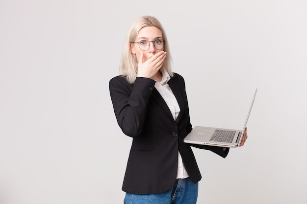 Blonde hübsche Frau, die den Mund mit den Händen mit einem schockierten bedeckt und einen Laptop hält